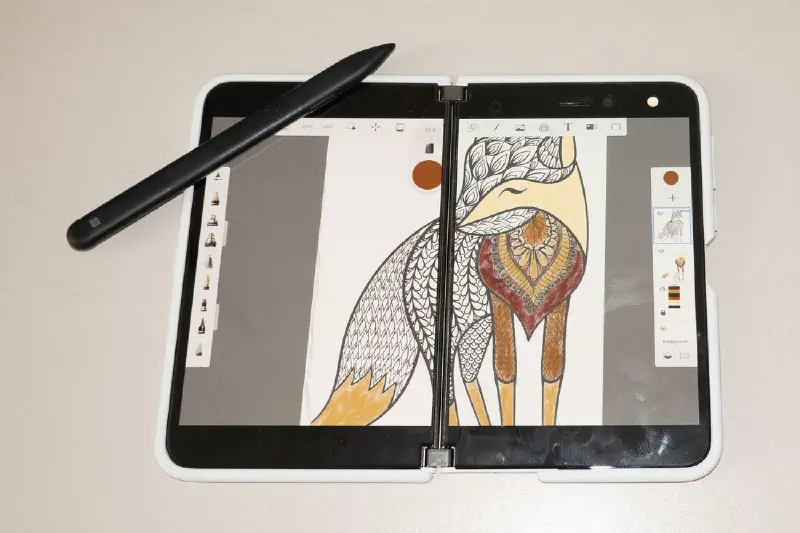 A Surface Duo foldable phone showing Sketchbook full screen and a Surface Pen sitting on the corner of the screen at an angle. The work shown on-screen is a partially complete coloring page.